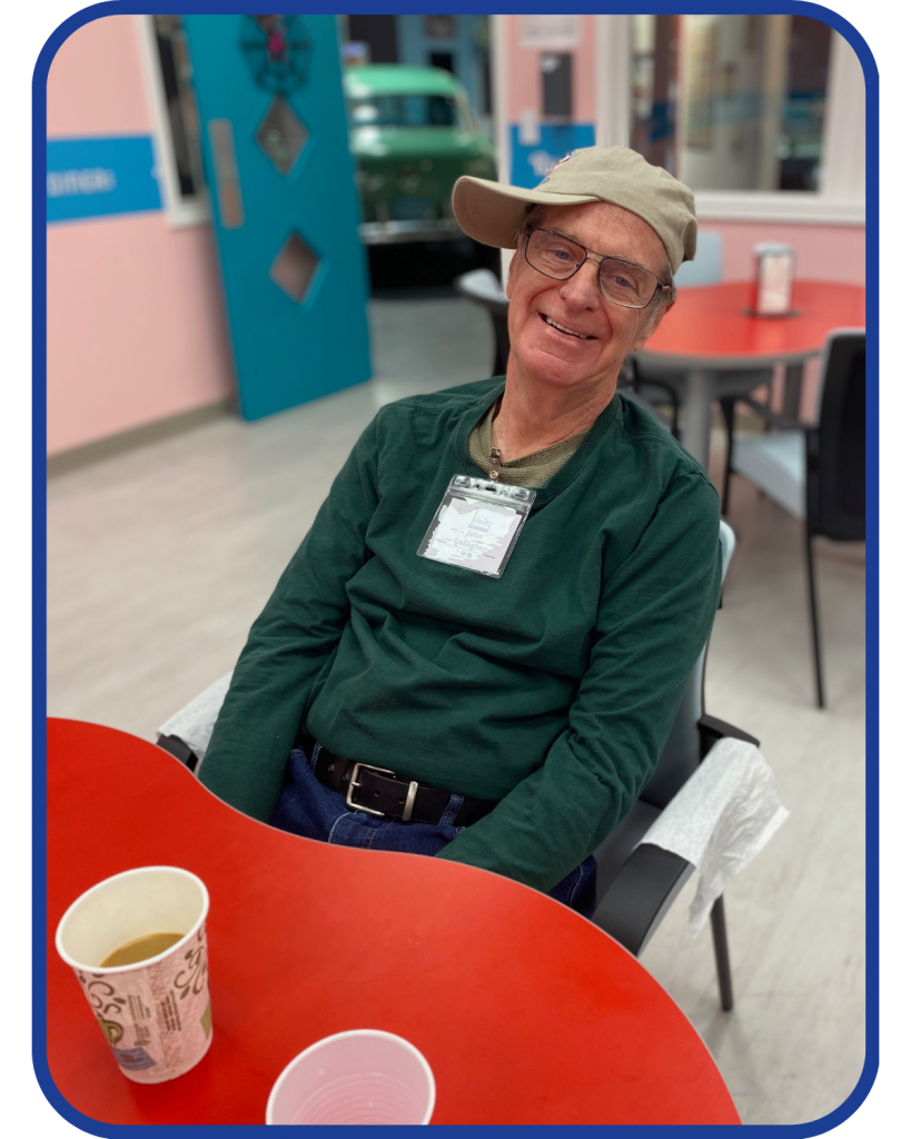 An olde adult sits at a red table with a cup of coffee. He is wearing a green shirt and a tan baseball cap cocked to the side. 