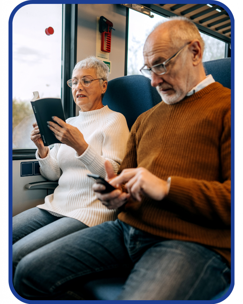 An older woman and man traveling by train. She is reading a book. He is looking at his phone. 