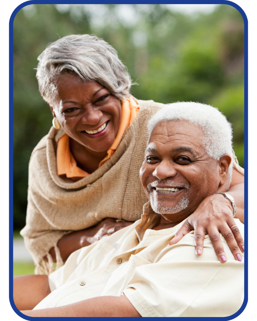 An older african american couple. The woman is leaning over the shoulder of the man and they are both smiling and looking at the camera.
