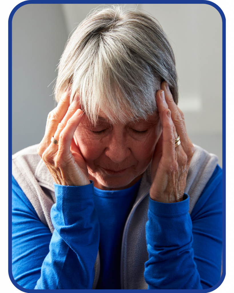 older adult female with white hair presses fingers to her temples appears stressed wearing a blue long sleeved shirt
