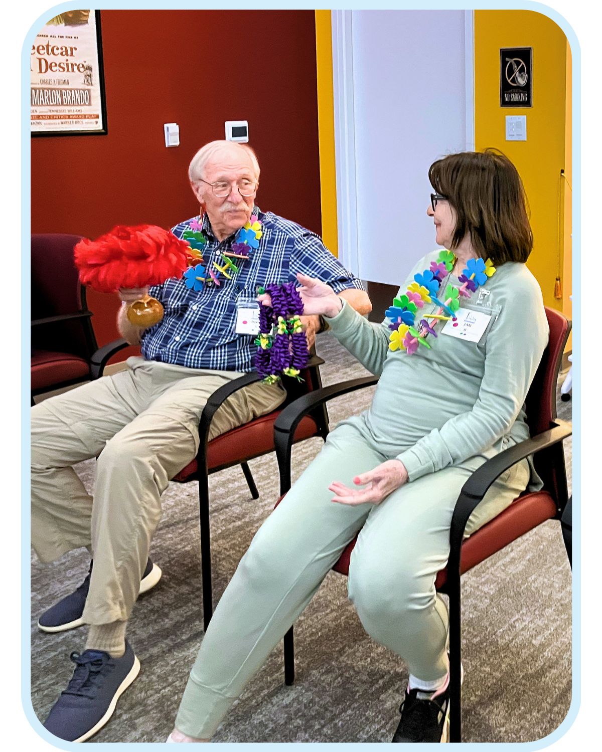 Two adults wearing colorful flower leis sit together and converse during an activity at Town Square NW Austin.
