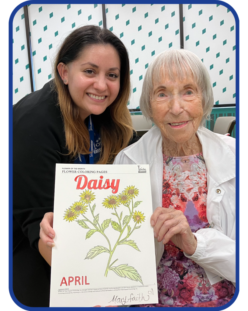 older female adult holds up a coloring page while her caregiver smiles beside her