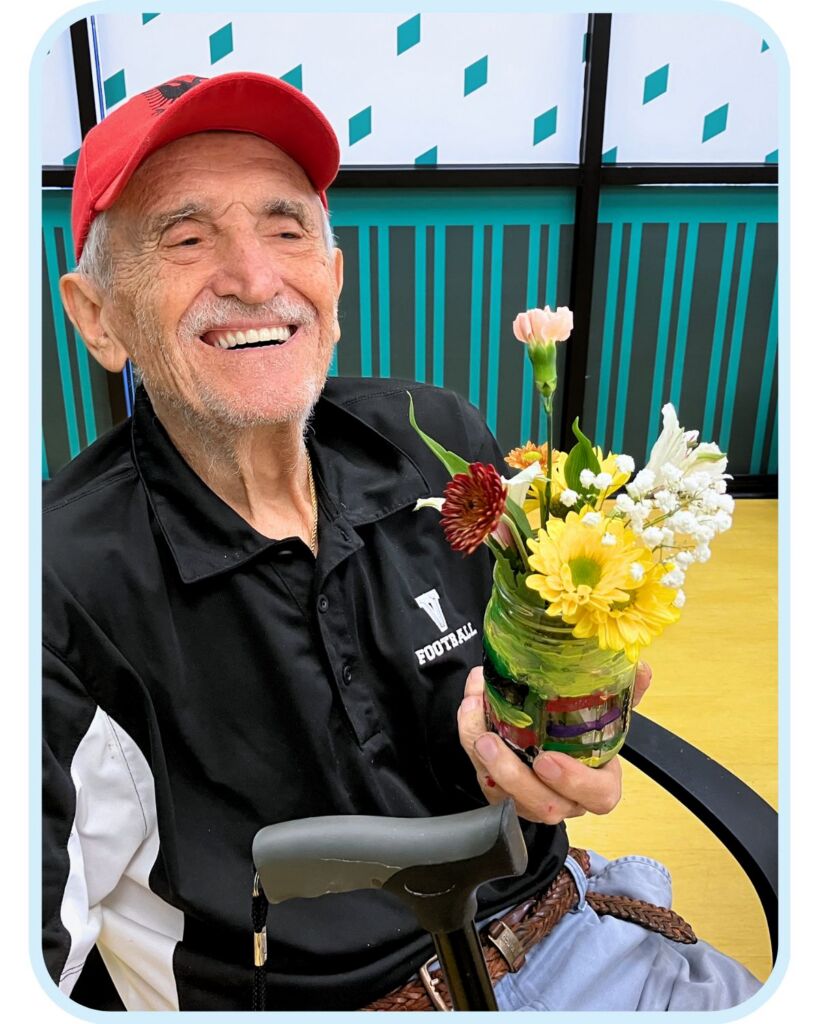 Smiling older man at Town Square NW Austin holding a small flower arrangement during a creative activity.
