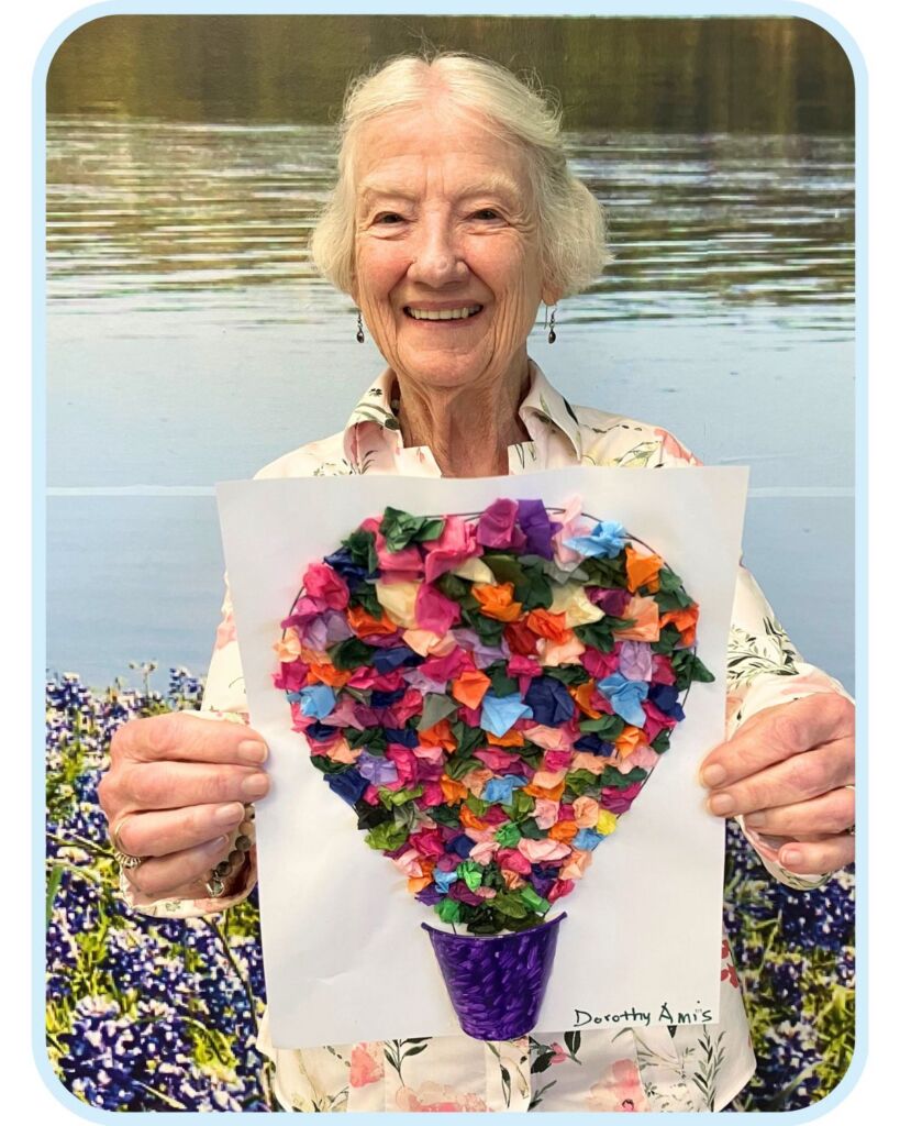 Smiling older woman at Town Square NW Austin holding a colorful tissue paper flower craft during an art activity.