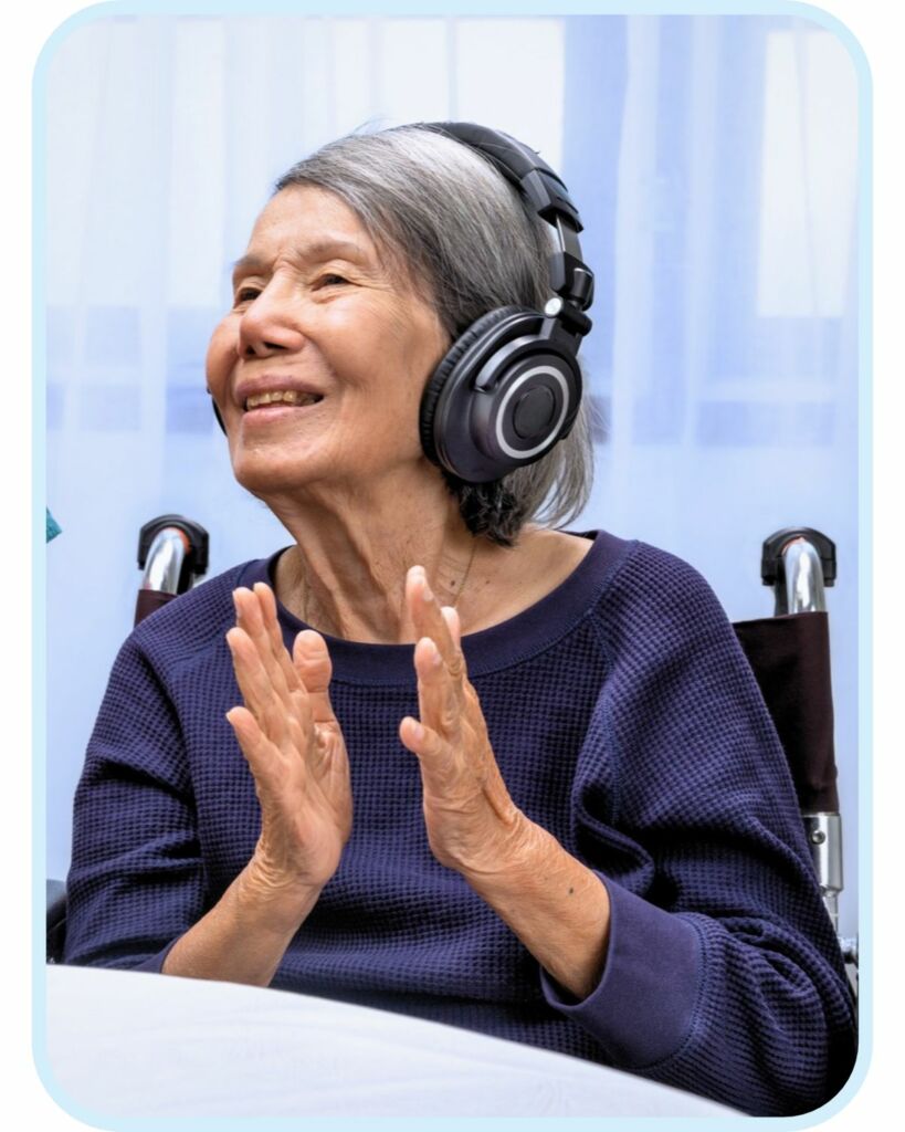 An older woman seated in a wheelchair smiles and claps while wearing over-ear headphones.